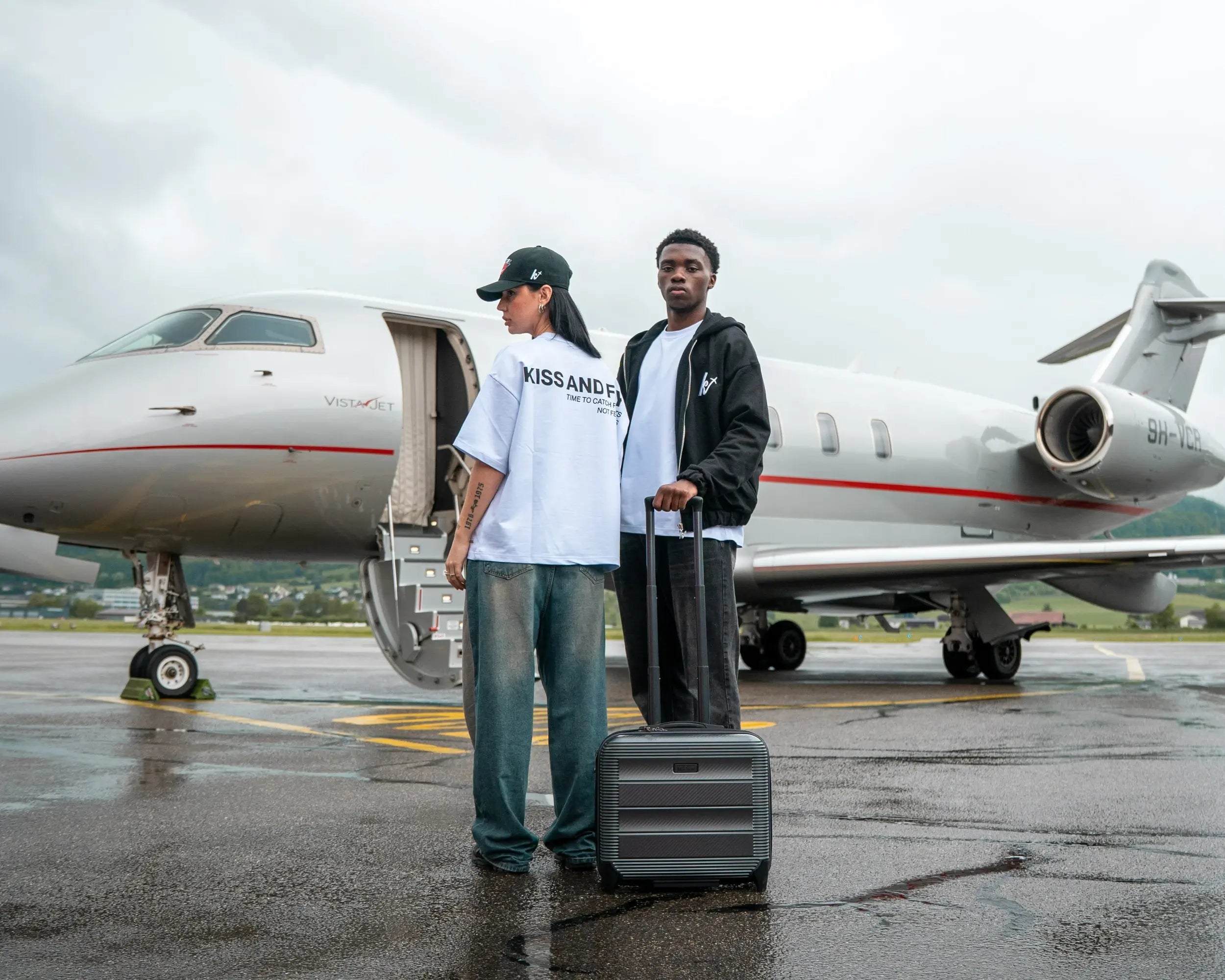 Two models wearing Kiss and Fly streetwear standing in front of a private jet. Female model in oversized white t-shirt with back print and cap, male model in black hoodie with suitcase. Luxury travel fashion scene.