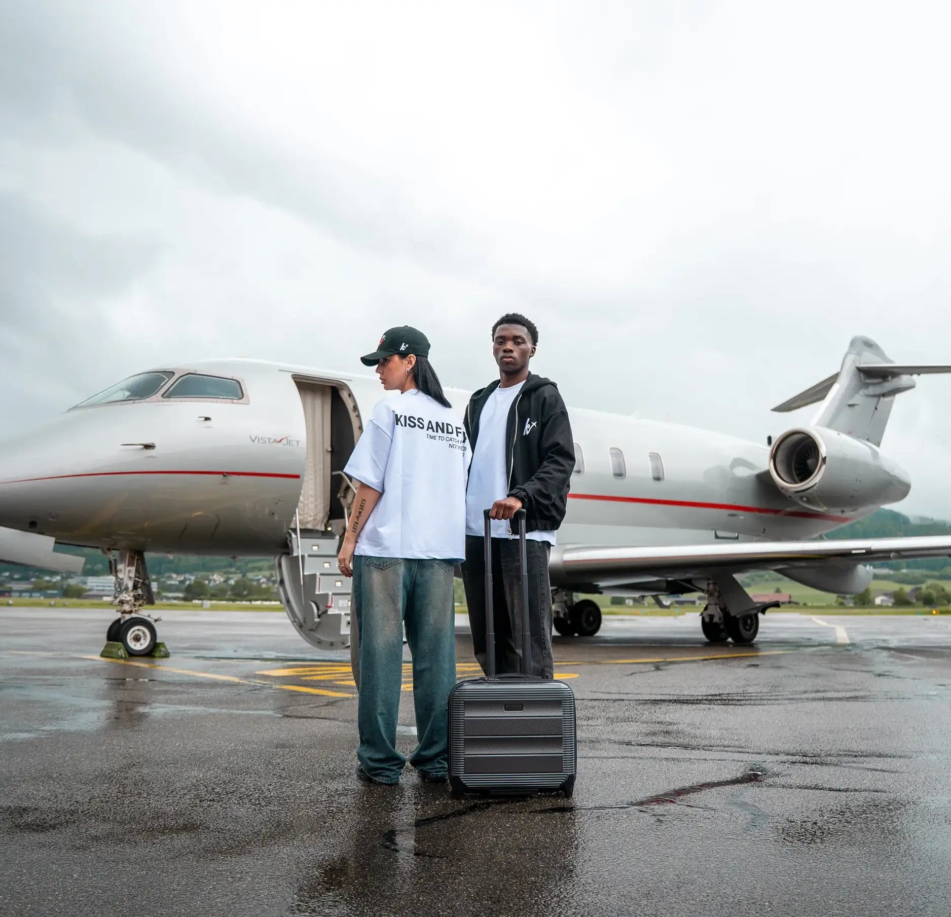 Two models wearing Kiss and Fly streetwear standing in front of a private jet. Female model in oversized white t-shirt with back print and cap, male model in black hoodie with suitcase. Luxury travel fashion scene.
